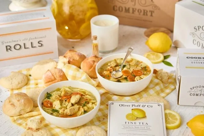 Two bowls of chicken and vegetable soup with carrots and herbs, styled on a yellow checkered cloth with bread rolls, lemon slices, and tabletop décor in the background.