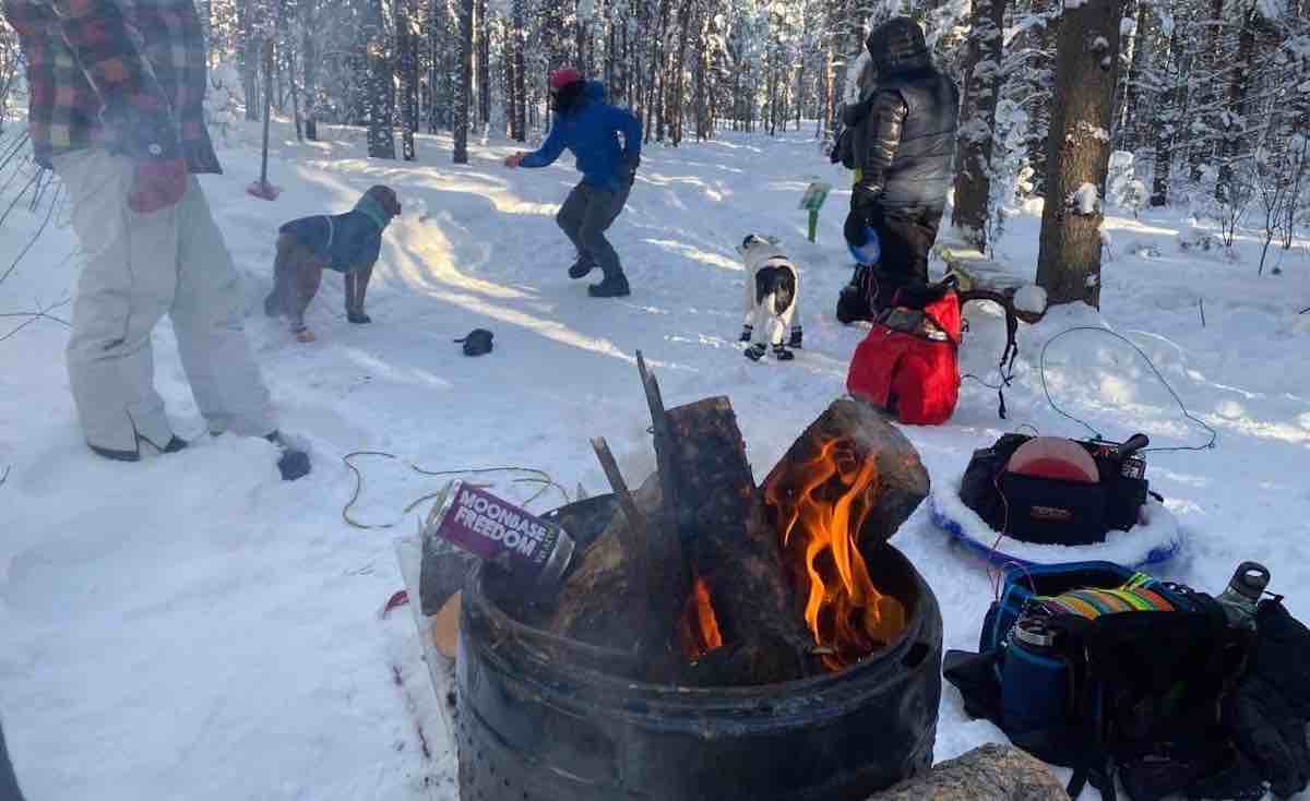 People playing disc golf in snow with barrel fire behind tee