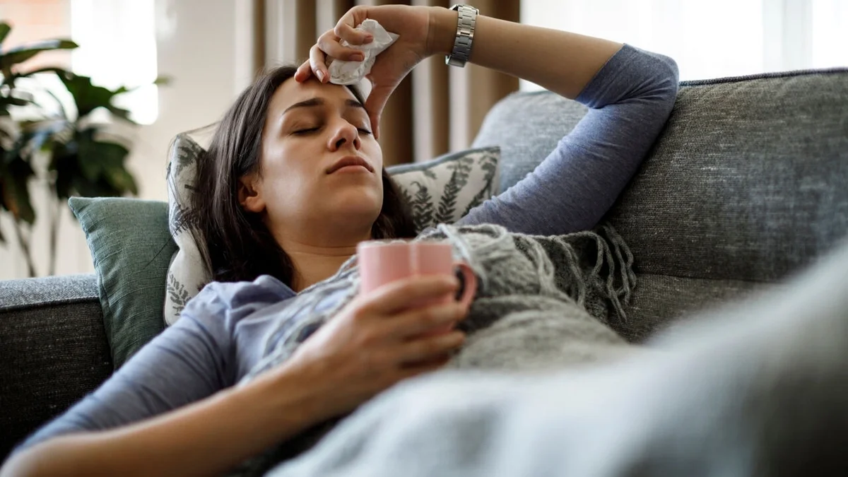 Person resting on a couch under a blanket, holding a mug while placing a cool cloth on their forehead.