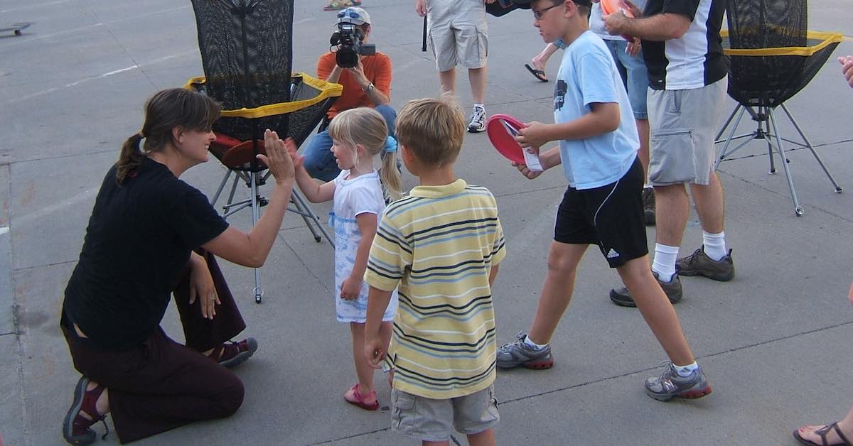A woman in a paved area with a group of kids and disc golf equipment. The woman is high fiving a young girl.