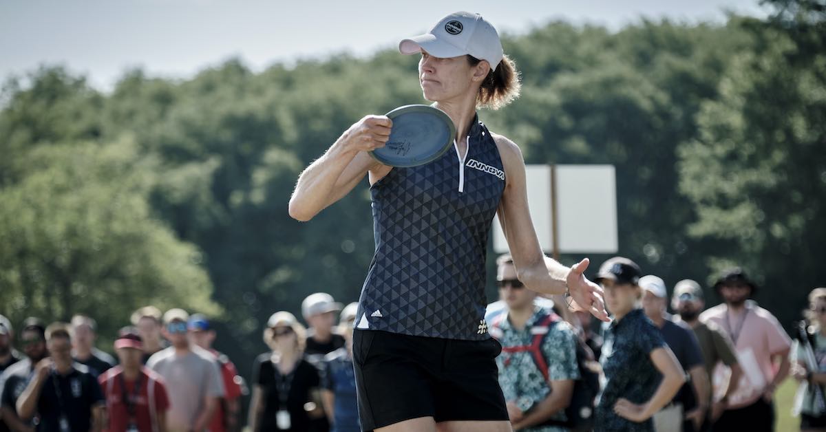 A woman in a baseball cap and athletic clothes prepares to throw a disc golf disc in front of a crowd