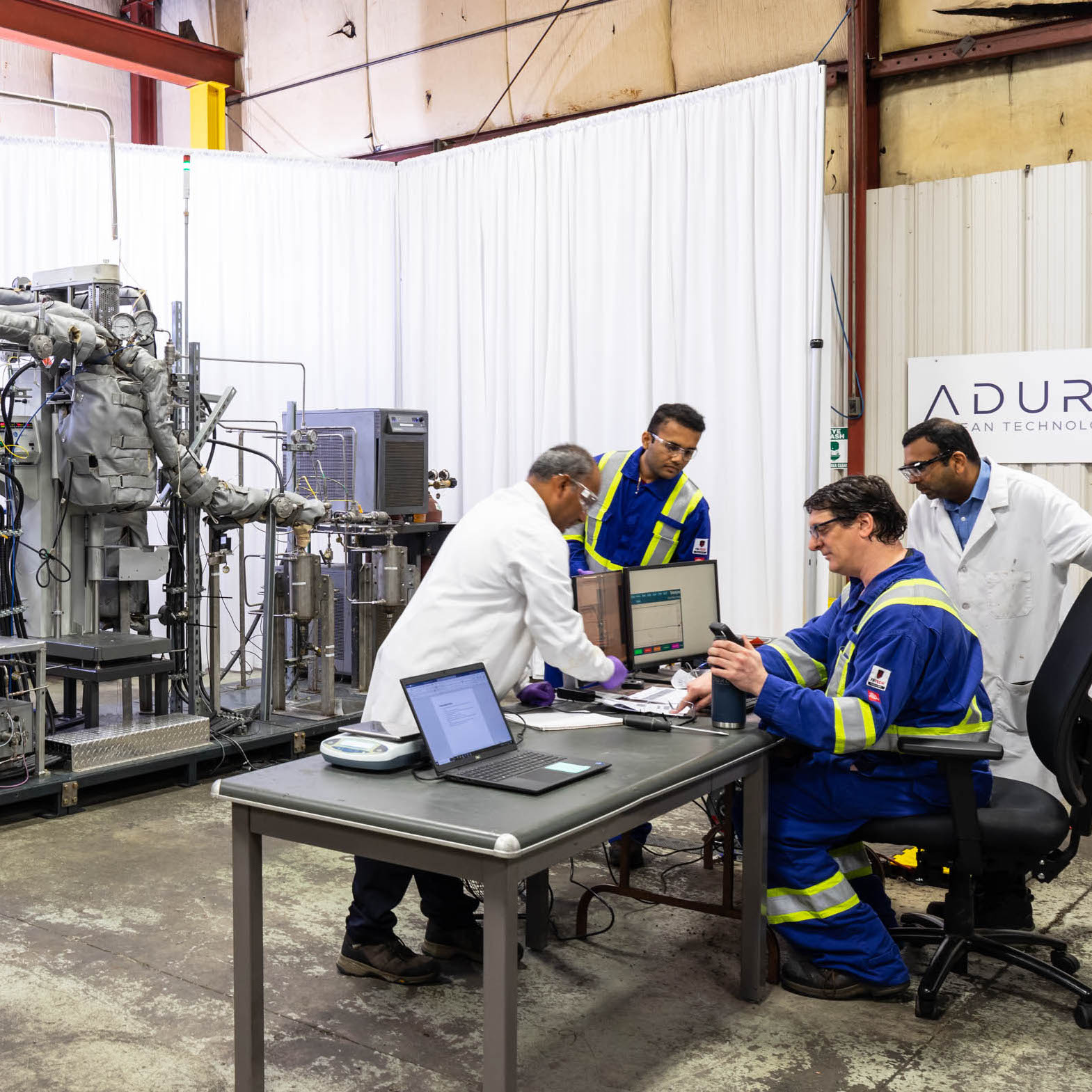 A group of scientists gather around a table with machinery in the background