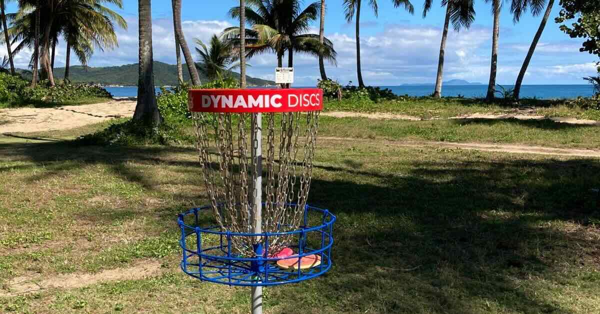 A red, white, and blue disc golf basket in front of a tropical beach
