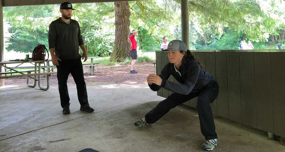 Trainer in a ball cap looks on as a female athlete performs warm up stretches