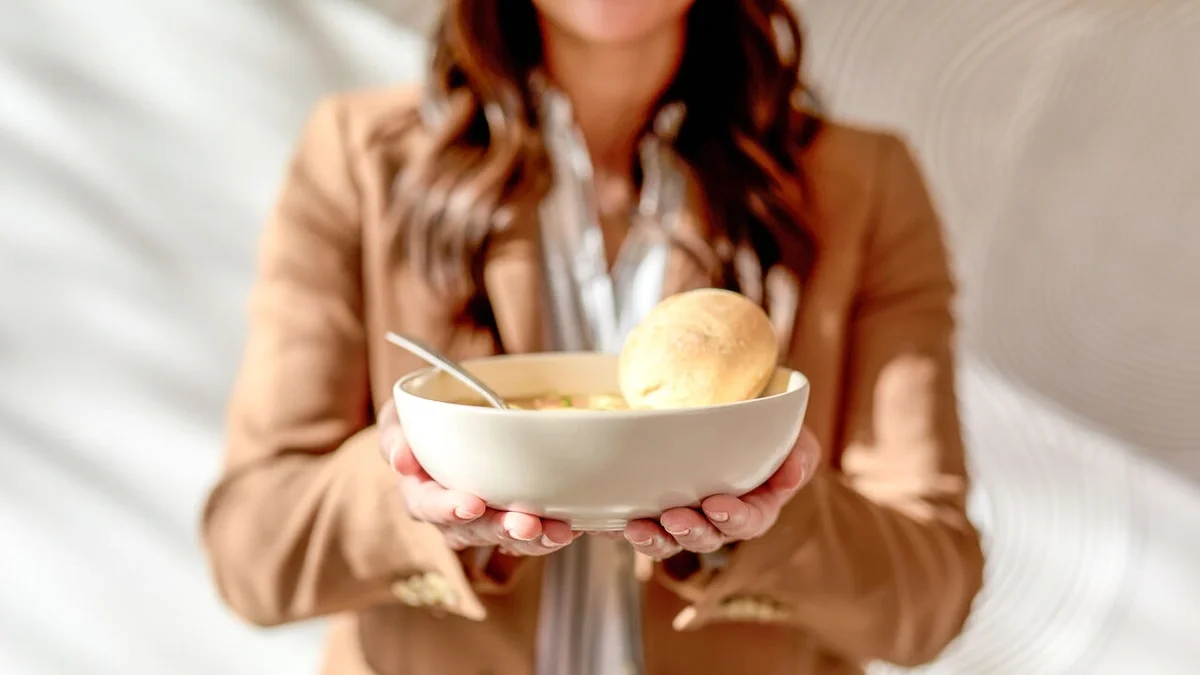 Person holding out a bowl of soup topped with a bread roll, offering it toward the viewer.
