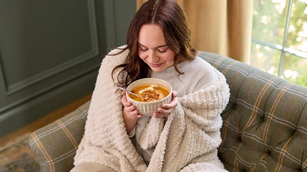 Woman wrapped in a cozy throw blanket, smiling as she holds a warm bowl of tomato soup while sitting in a plaid armchair.