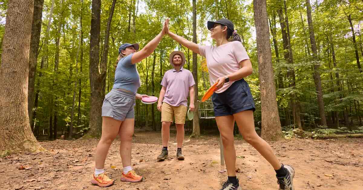 Two women high five with disc golf discs in their hand while another player smiles near a disc golf basket