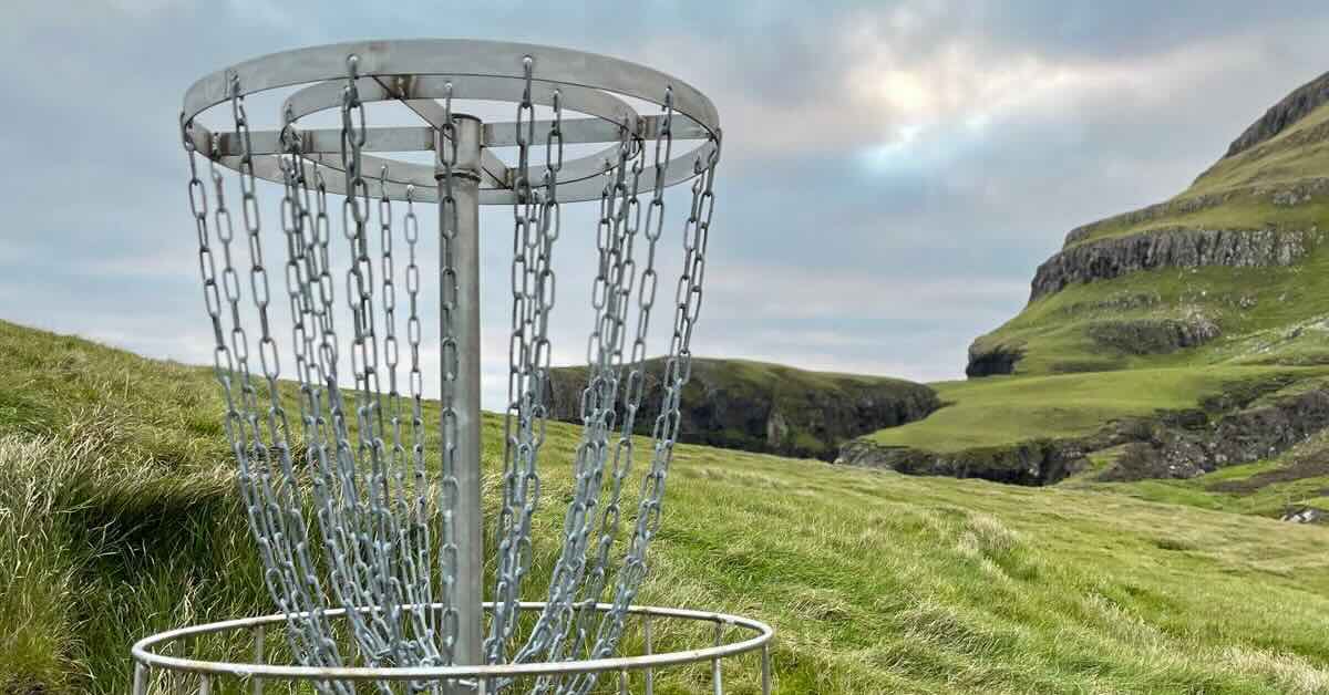 A metal disc golf basket in a grassy area with an exposed rock face on the right