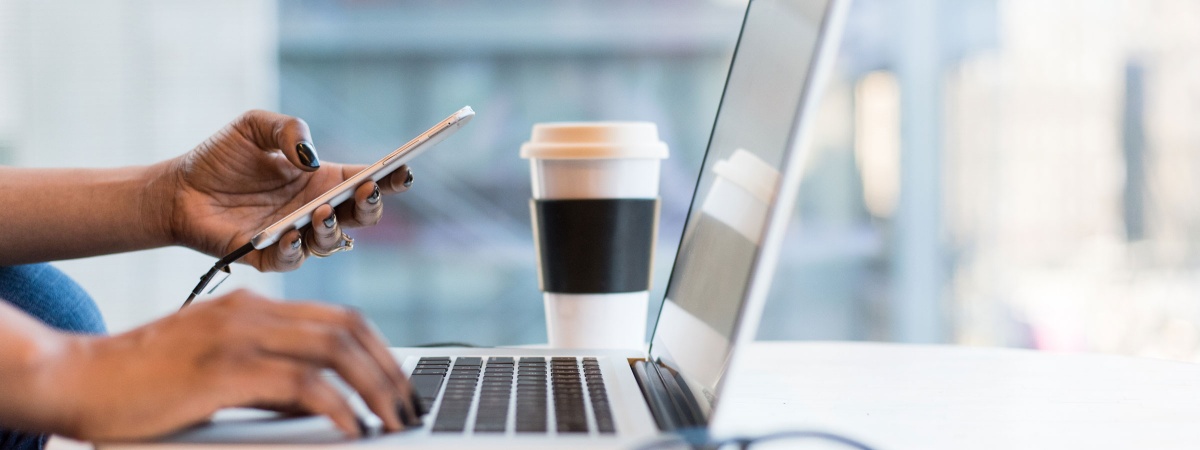 Closeup of woman's hands using laptop and cell phone, with coffee cup in background