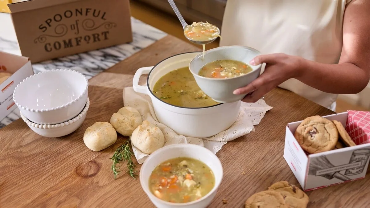 Person ladling chicken soup with carrots and herbs from a white pot into a bowl, surrounded by fresh rolls, cookies, and Spoonful of Comfort packaging on a wooden table.