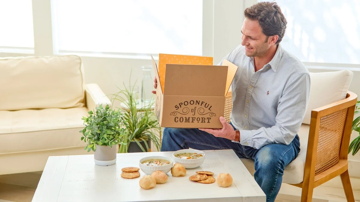 Man sitting in a bright living room, smiling as he opens a Spoonful of Comfort gift box, with bowls of soup, rolls, cookies, and potted plants on the coffee table.