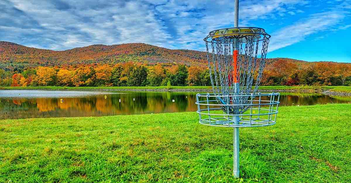 A disc golf basket in front of a body of water and mountains with trees in fall colors