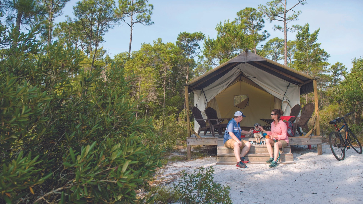 Two people sit on the steps of a tent set up in a wooded area. The two are relaxing and surrounded by nature on a sunny day.