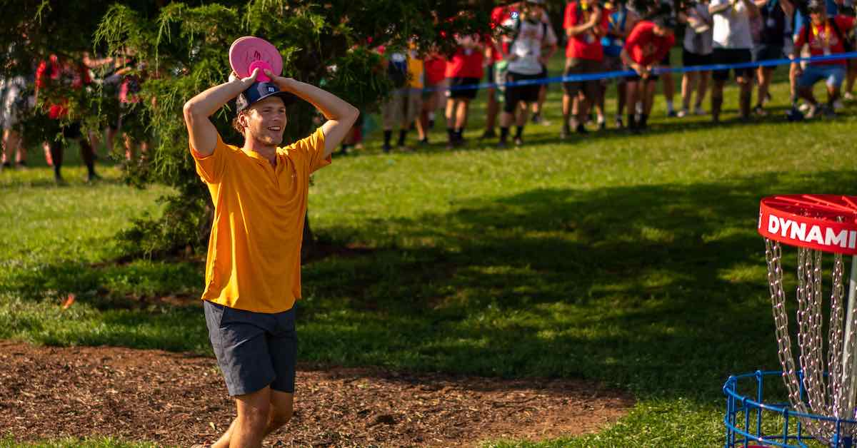 Young man in organge shirt and ball cap smiles in wonder and holds disc above his head as he approaches a basket in front of a crowd