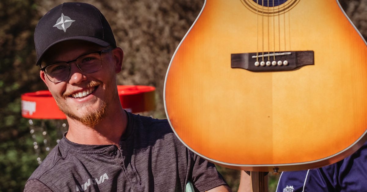 A young man with a ball cap and beard holds up a guitar and smiles with top of disc golf basket in background