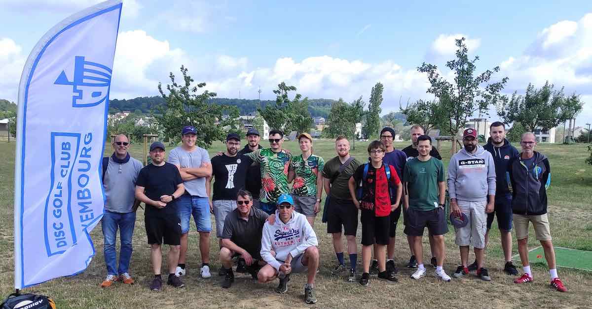 A group photo in a park area with young trees and a white flanner with "Disc Golf Club Luxembourg" in blue