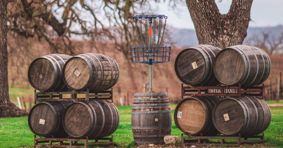 A blue disc golf basket on top of a vertical wine barrel with neatly-stacked wine barrels beside it