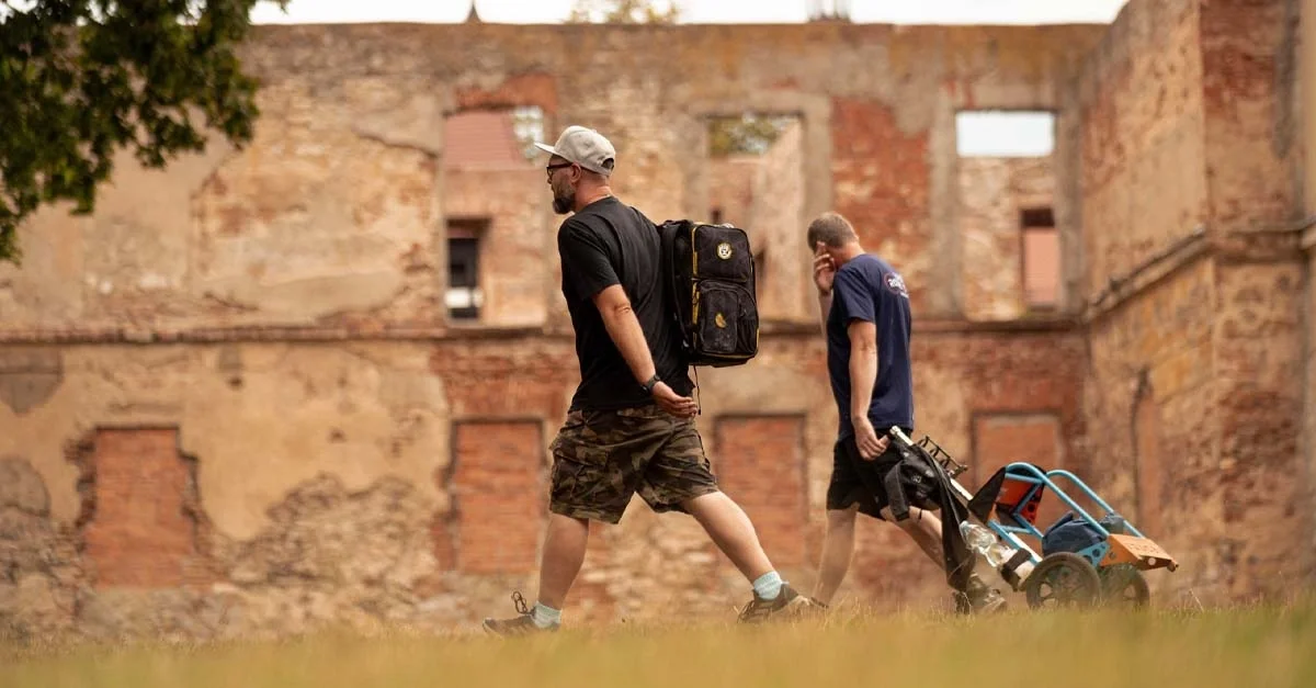 A man wearing a hat walks with a backpack, while another man pulls a cart, both in front of an antiquated building