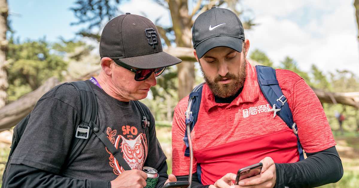 Two men outdoors in baseball caps look at one man's phone
