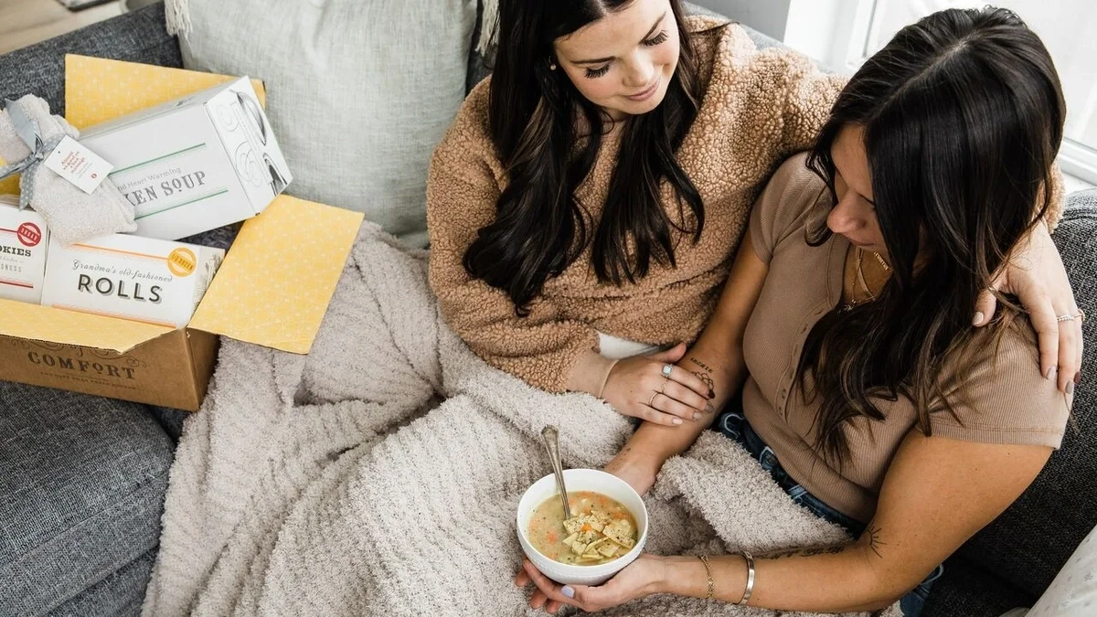 Overhead view of two women sitting together under a cozy blanket, with one offering comfort by resting a hand on the other's arm while a bowl of soup and an open Spoonful of Comfort gift box sit nearby.