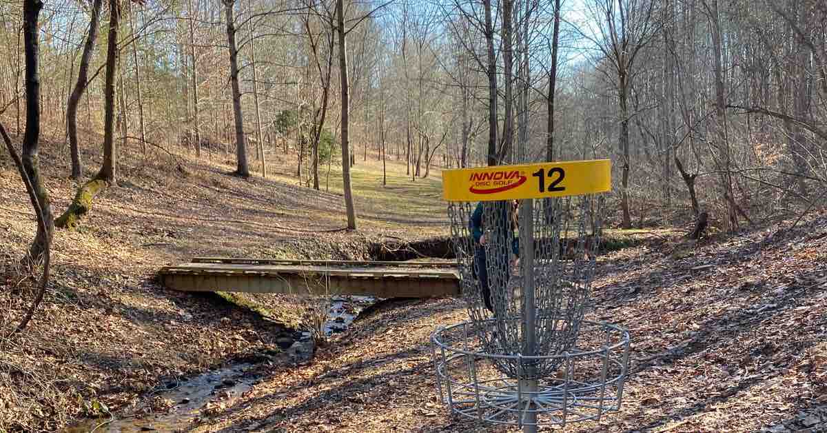 A disc golf basket with a yellow band near a stream in a wooded area