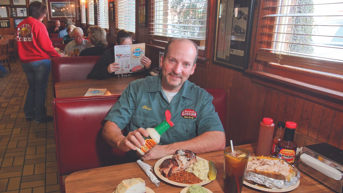 A man in a green shirt sits in a booth at a restaurant, holding a bottle of sauce over a plate of food. The table holds an iced tea, baked potato, side dishes, and sauce bottles.