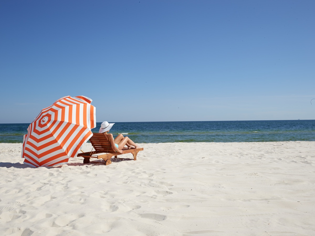 A beach umbrella sits on the sand next to a woman on a beach chair overlooking Alabama's Gulf Coast.