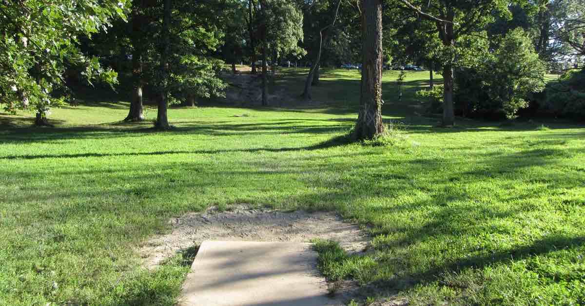 A concrete disc golf tee pad in a park area with mown grass and mature trees