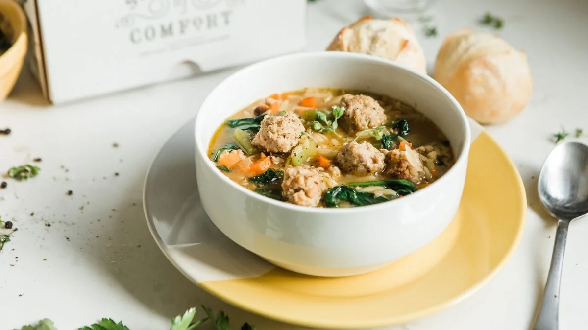 Close-up of a bowl of hearty soup with meatballs, carrots, leafy greens, and herbs, served with fresh rolls and Spoonful of Comfort packaging in the background.