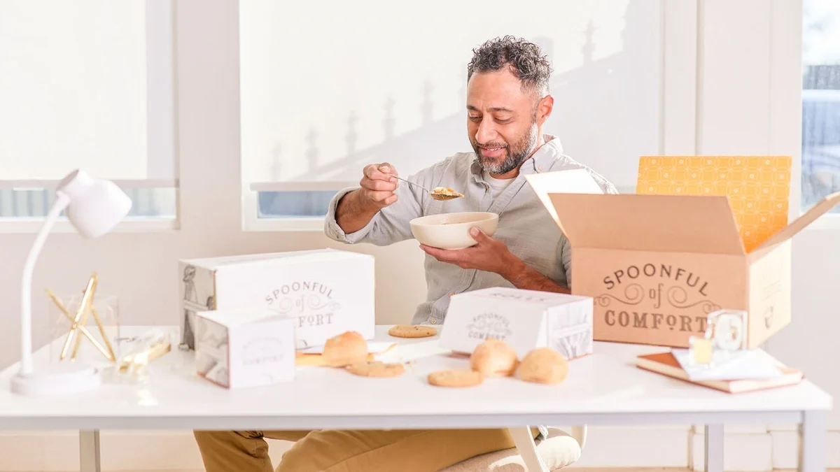 Man sitting at a desk enjoying a bowl of soup beside an open Spoonful of Comfort gift box, with packaged soup, rolls, cookies, and a handwritten note spread across the table.