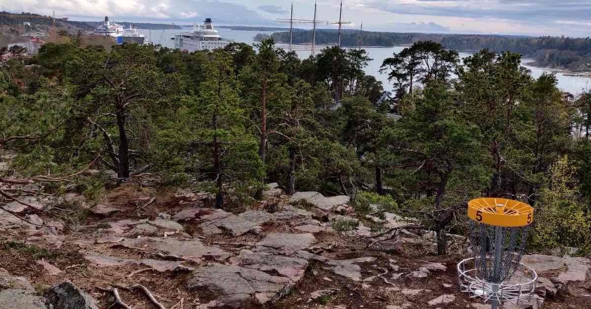 A disc golf basket on a rocky landcape with water and large ships in the distance