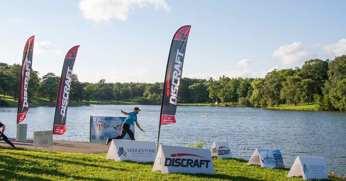 A player tees off, throwing a disc over a large lake with feather banners waving in the wind