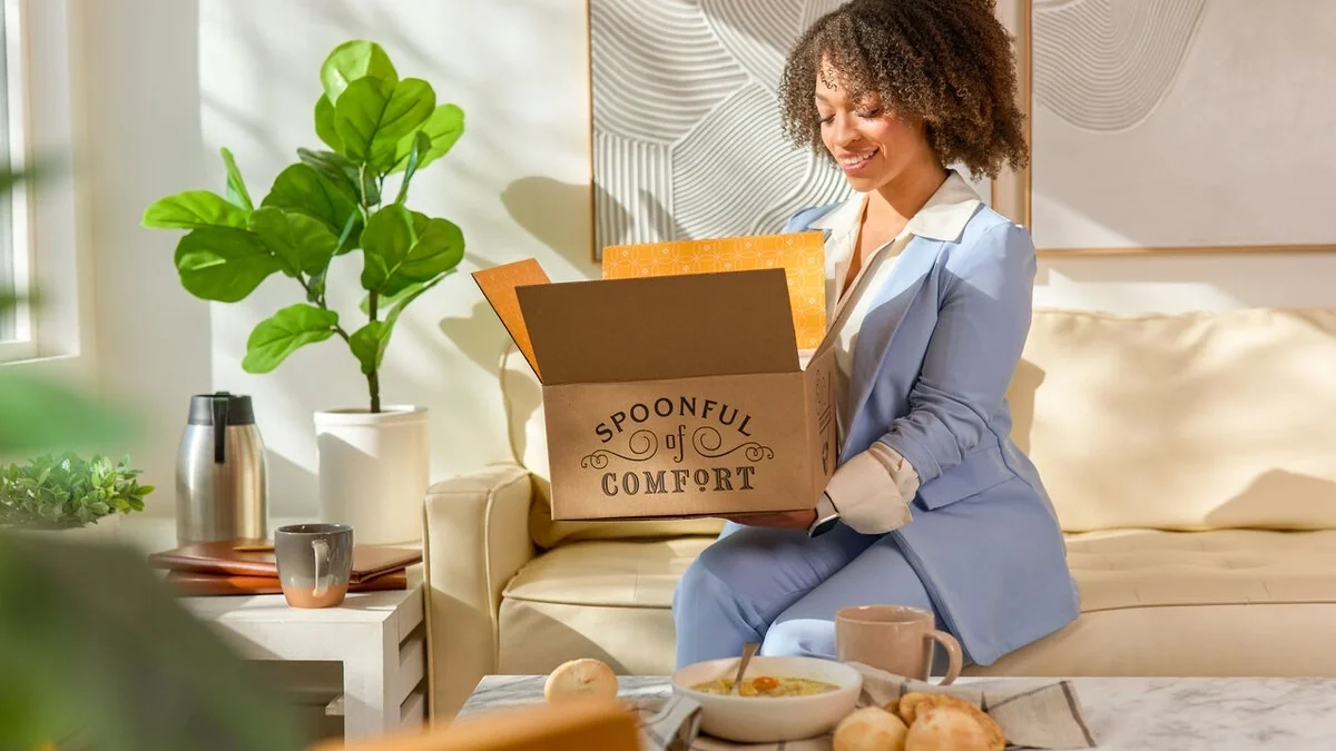 Woman sitting on a couch smiling as she opens a Spoonful of Comfort gift box, with a bowl of soup, rolls, and a mug on the coffee table in front of her.