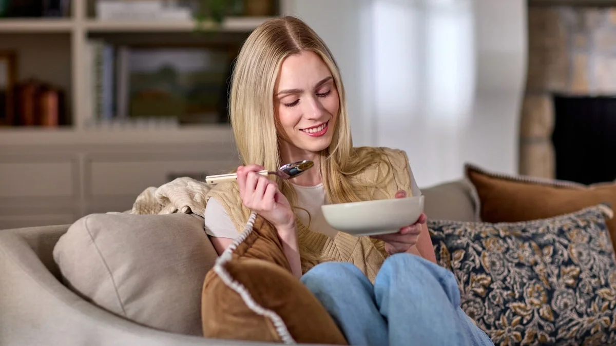 Young woman sitting on a couch, smiling while holding a bowl of soup and a spoon in a cozy living room.