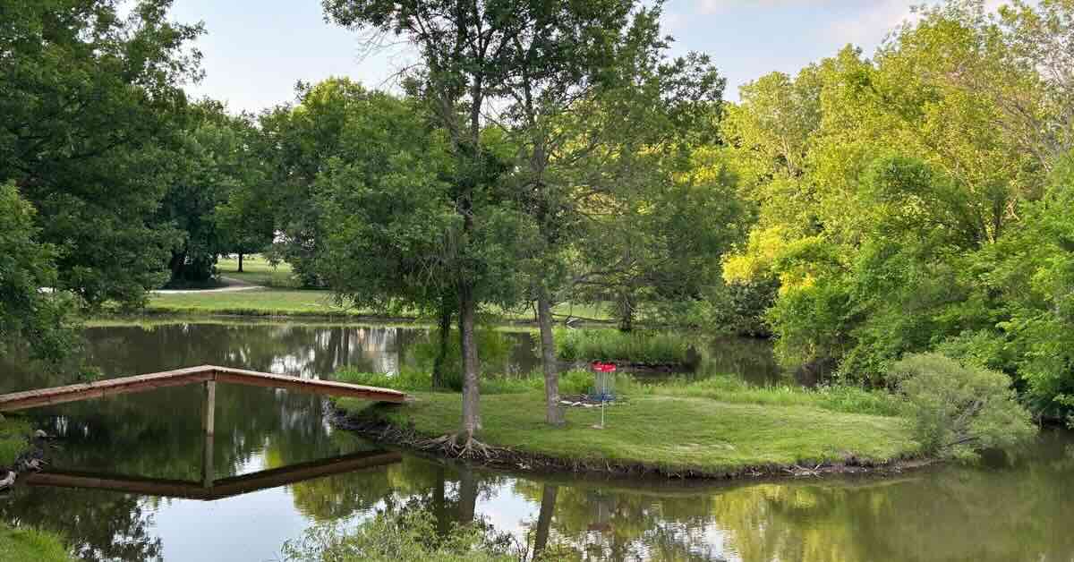 A blue and red disc golf basket on an island in a large pond with a bridge going to it