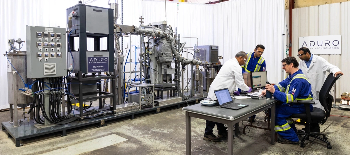 Lab technicians around a table looking at test results. Beside them is Aduro's plastics reactor, a large grey machine with several components.