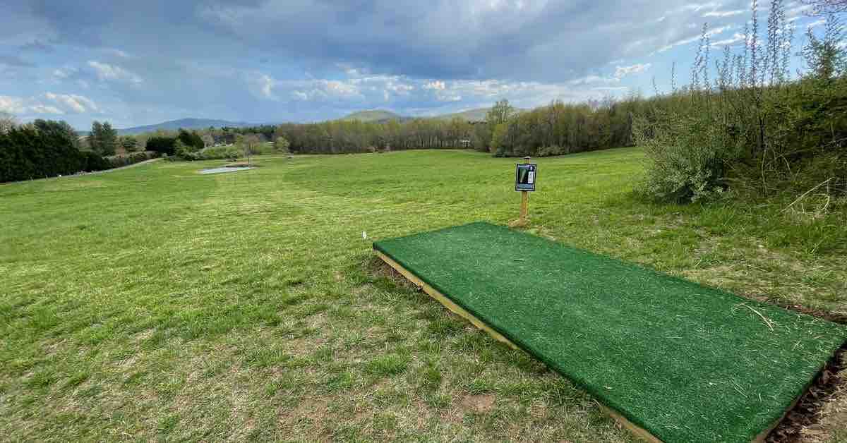 A turf disc golf tee pad leads to a view of well-mown field on a cloudy day
