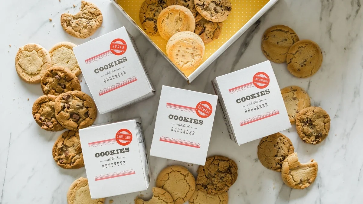 Overhead view of four boxes of assorted cookies surrounded by chocolate chip, oatmeal, and sugar cookies on a marble surface, with an open gift box in the background.