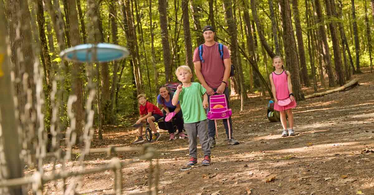 A small child throwing a disc into a disc golf basket as the fmaily looks on