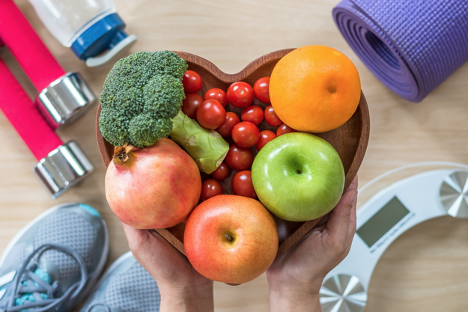 Woman’s hands holding heart shaped wooden bowl of fruit and vegetables with fitness equipment in the background.