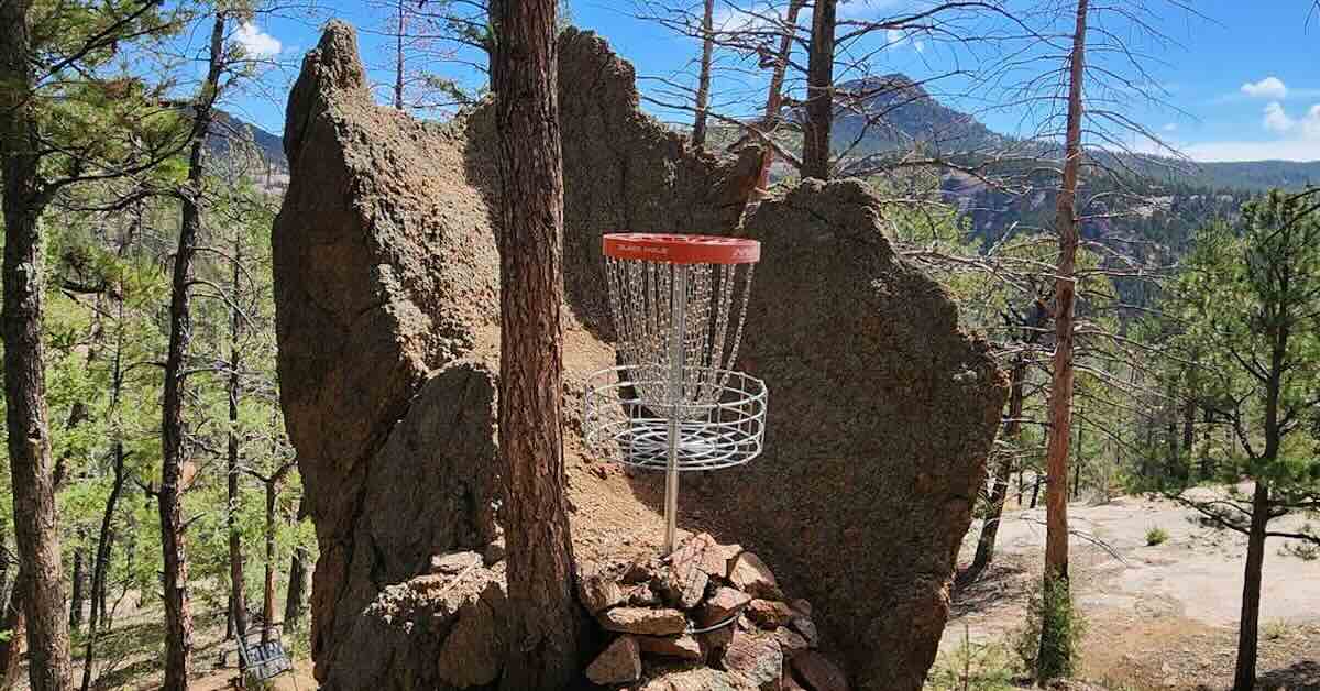 A red-banded disc golf basket in a strange rck formation with scattered trees and mountains beyond