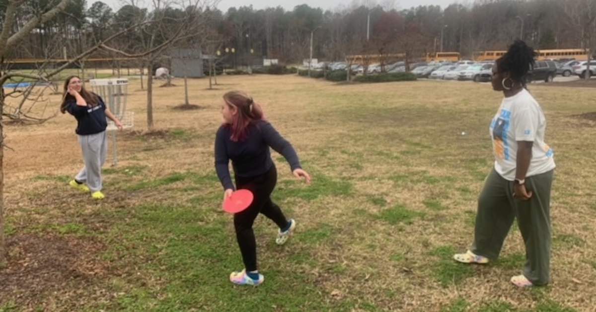 Three young girls outside on a chilly, gray day playing disc golf on a school campus