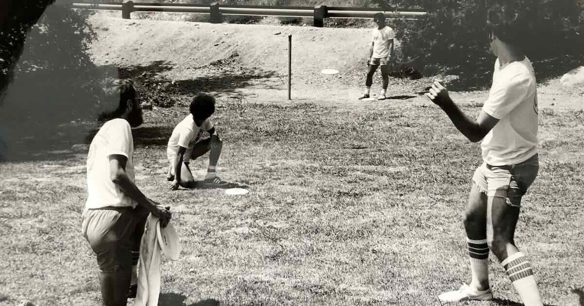 Men in 70s athletic outfits in a black and white photo. One throws a Frisbee at a pole