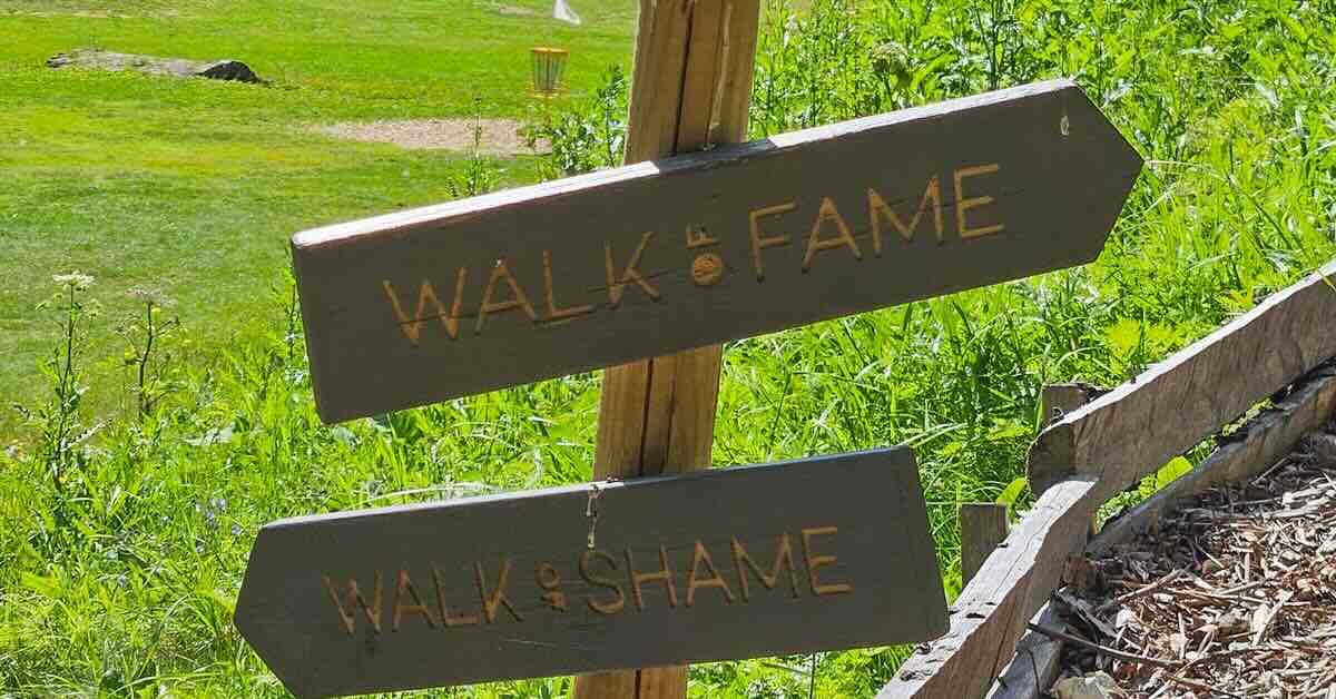 Two directional signs at a disc golf course saying "walk of shame" and "walk of fame"
