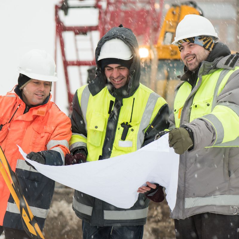 Three people in construction workwear for winter looking at a blueprint while snow is falling on them