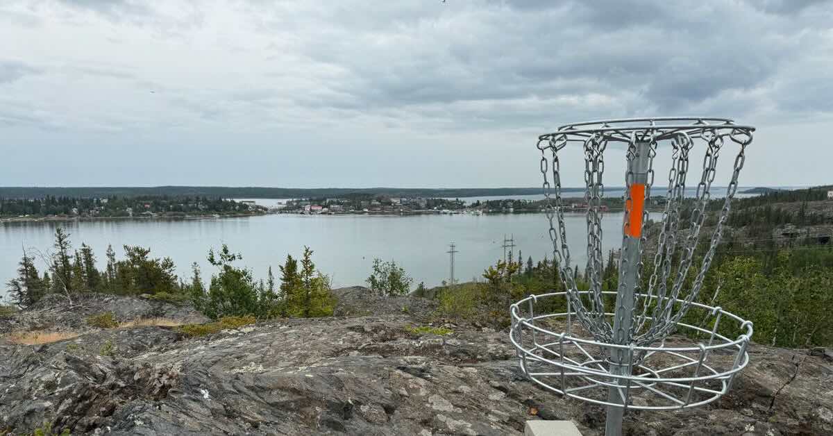A disc golf basket on exposed rock with a view of open water and homes built on land in the water beyond