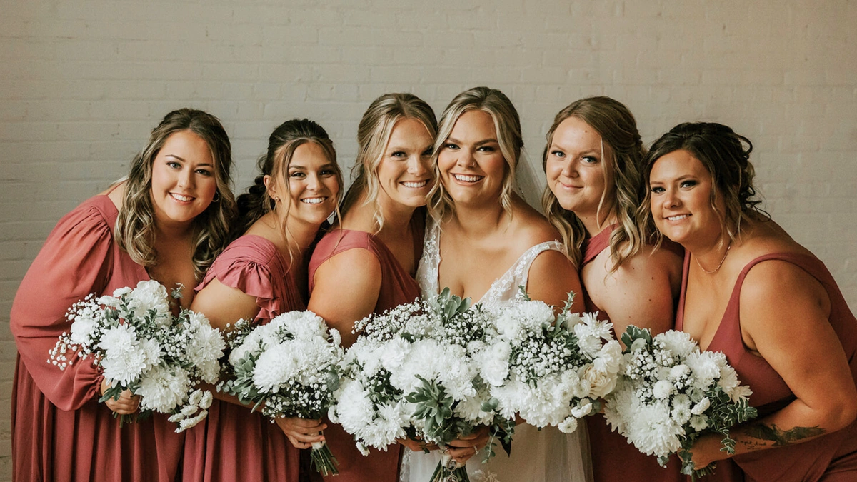 Smiling bride and bridesmaid holding neutral, white and green wedding bouquets with roses and baby's breath.